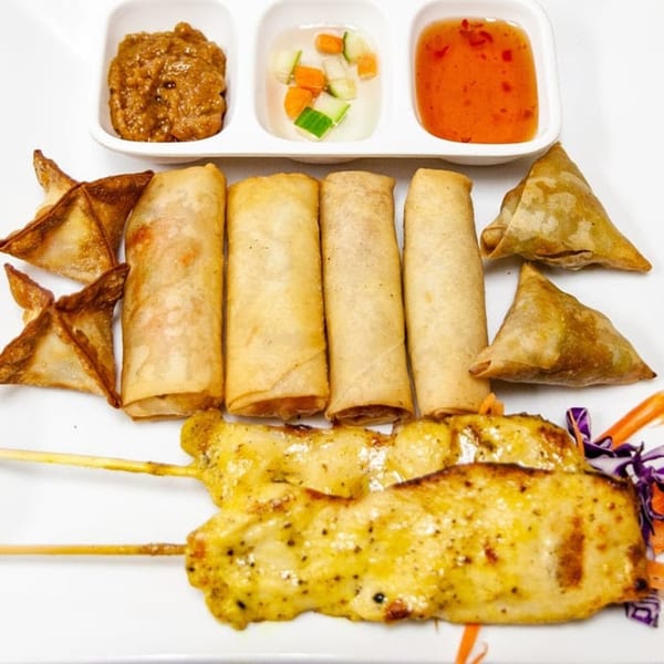 A Thai appetizer platter with spring rolls, crab rangoons, samosas, chicken satay, and dipping sauces.