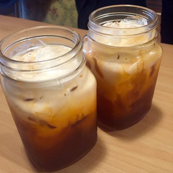 Two glasses of Thai iced tea with ice and cream on a light wooden table.